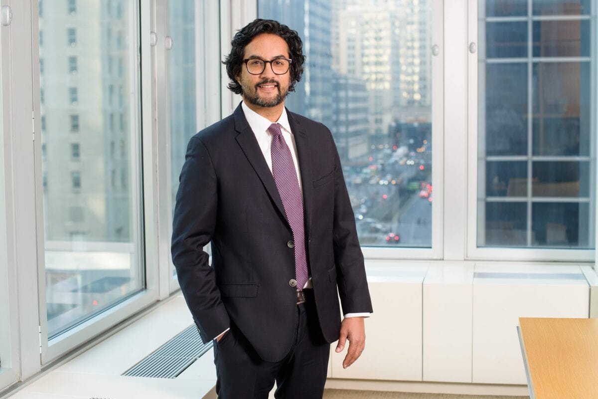 Headshot example featuring a man in a dark suit with a casual hand-in-pocket pose, framed by a high-rise office window grid overlooking an urban street scene.
