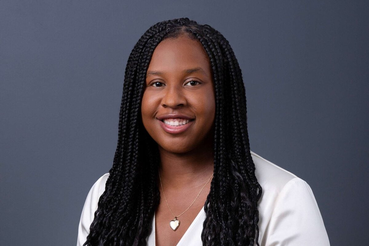 Headshot example featuring a professional woman with long braids and a white V-neck top, captured against a uniform, cool-toned blue-gray backdrop for a polished, modern look.