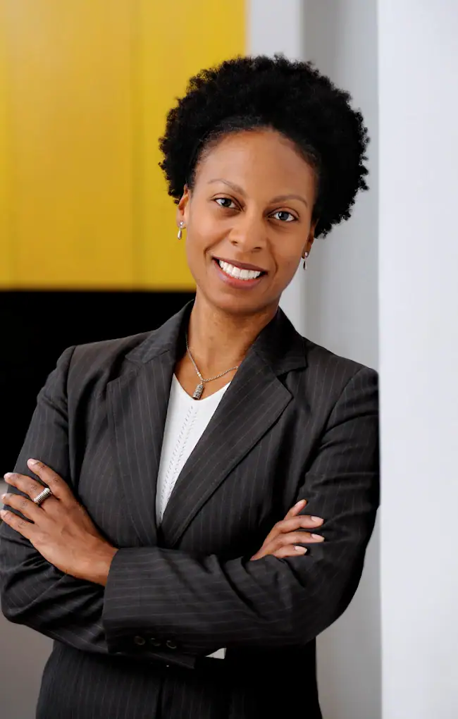 Headshot example featuring a woman in a dark pinstripe suit and white scoop-neck top with a relaxed lean against a white column, contrasted against a vibrant mustard-yellow background wall.