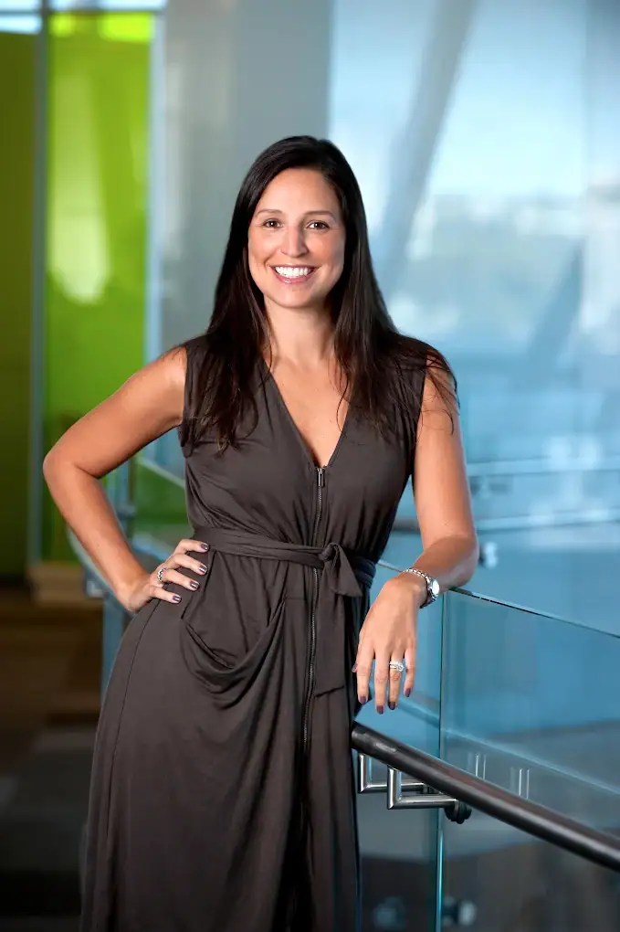 Headshot example featuring a woman in a modern dark brown dress with a unique leaning stance, set against a dynamic office backdrop with bright green and blue accents.
