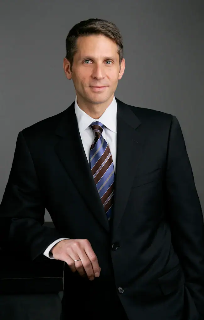 Headshot example featuring a male professional in a sharp black suit and diagonally striped blue and brown tie, leaning forward with controlled confidence against a neutral gray studio background.