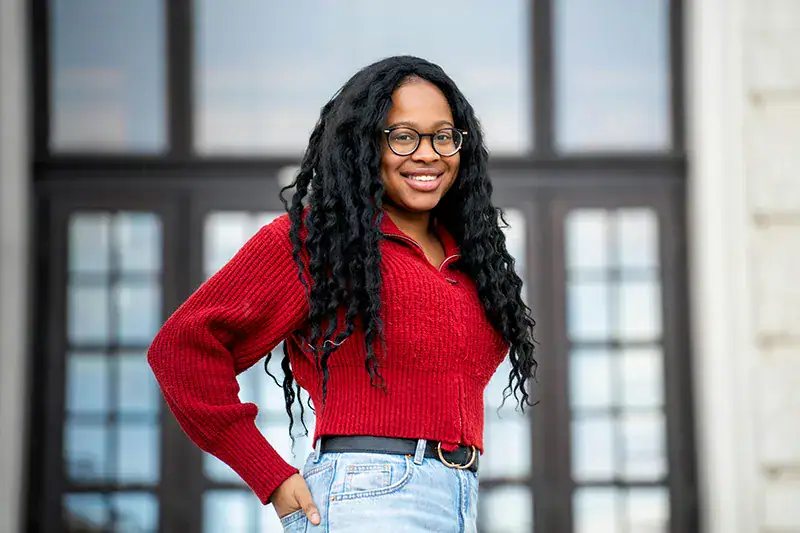 Headshot example featuring a woman with long dark hair in a bold red sweater and scholarly glasses, standing in an environmental pose before large architectural glass-paneled doors.