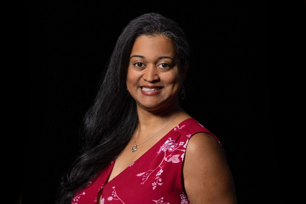 Headshot example featuring a woman in vibrant red patterned attire with a broad smile, utilizing strategic dimensional lighting to emerge from a low-key, shadowless black background.