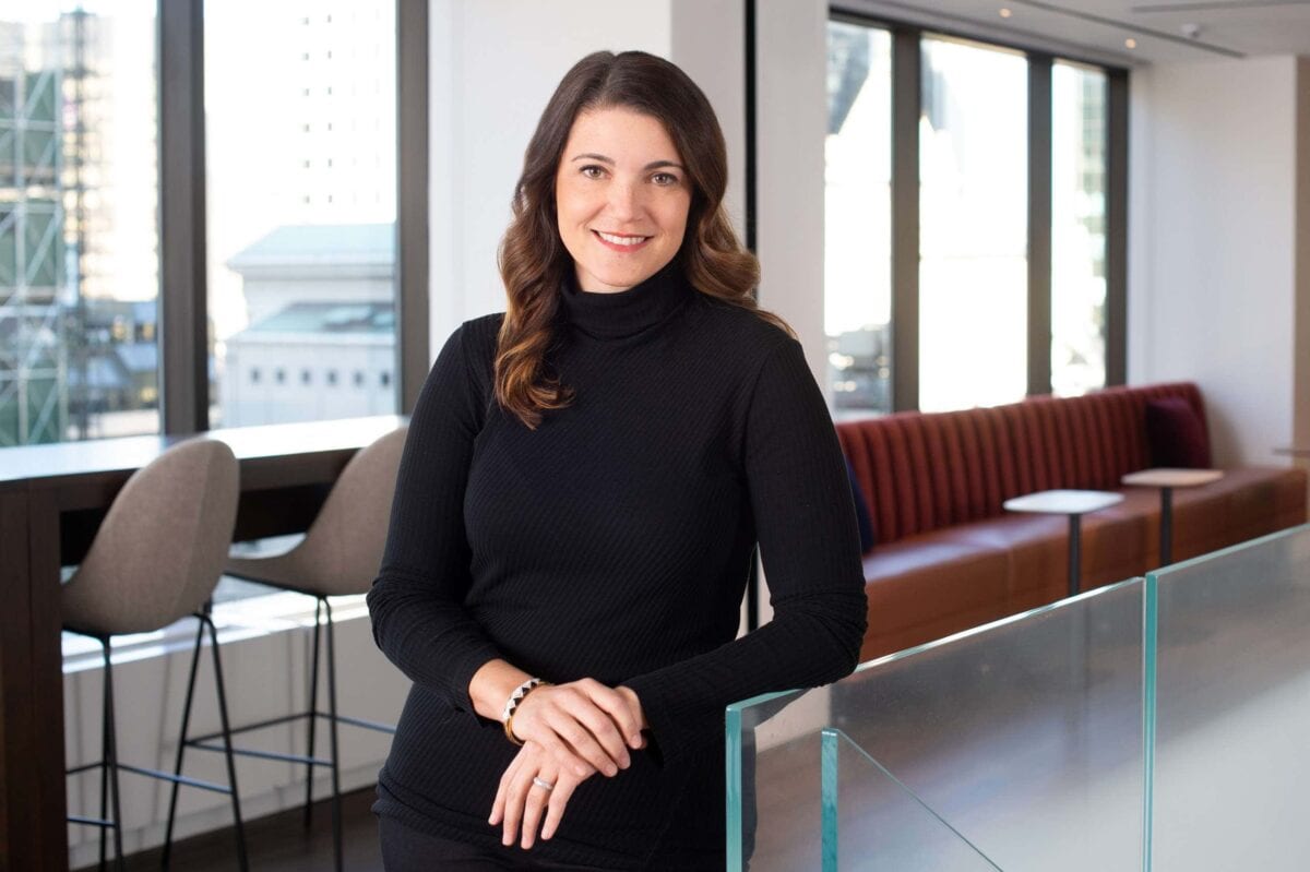 Headshot example featuring a woman in a black turtleneck leaning against a glass counter in a high-end office interior with abundant natural light.