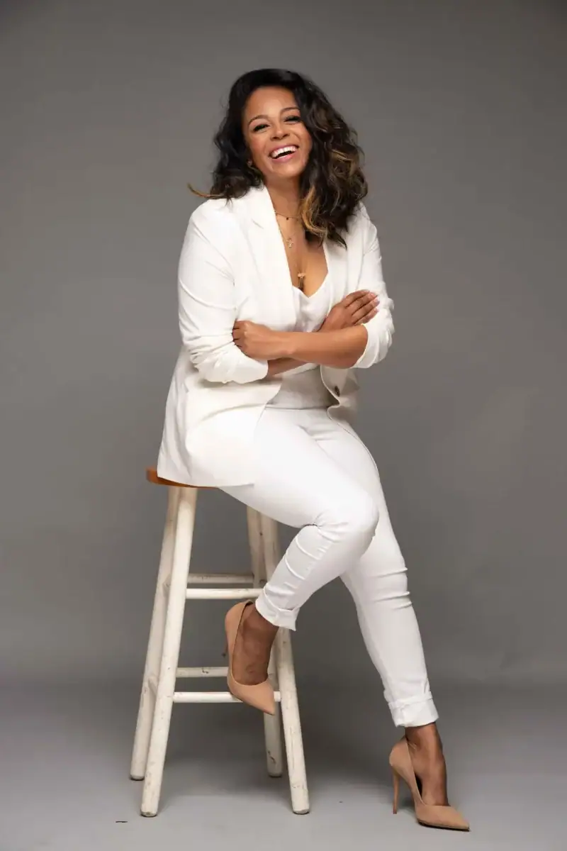 Headshot example featuring a person in an all-white outfit seated on a stool against a neutral gray studio background. A wide smile, crossed arms, and lively posture give the image an energetic, confident, polished feel.