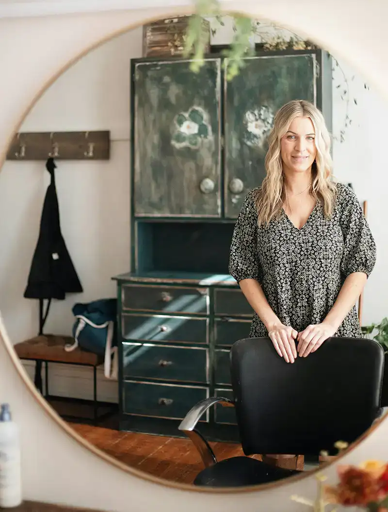 Headshot example featuring a professional stylist leaning on a salon chair, captured through a circular mirror frame within a rustic studio featuring a weathered green hutch and warm textures.
