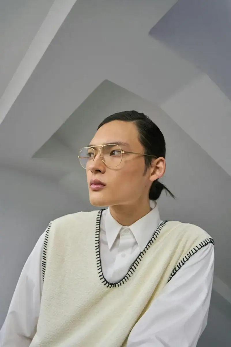 Headshot example featuring a person with hair pulled back, wearing thin gold glasses, a white collared shirt, and a cream knit vest with dark trim. Angular ceiling lines in the gray architectural background and the sideward gaze give the image a modern, editorial feel.
