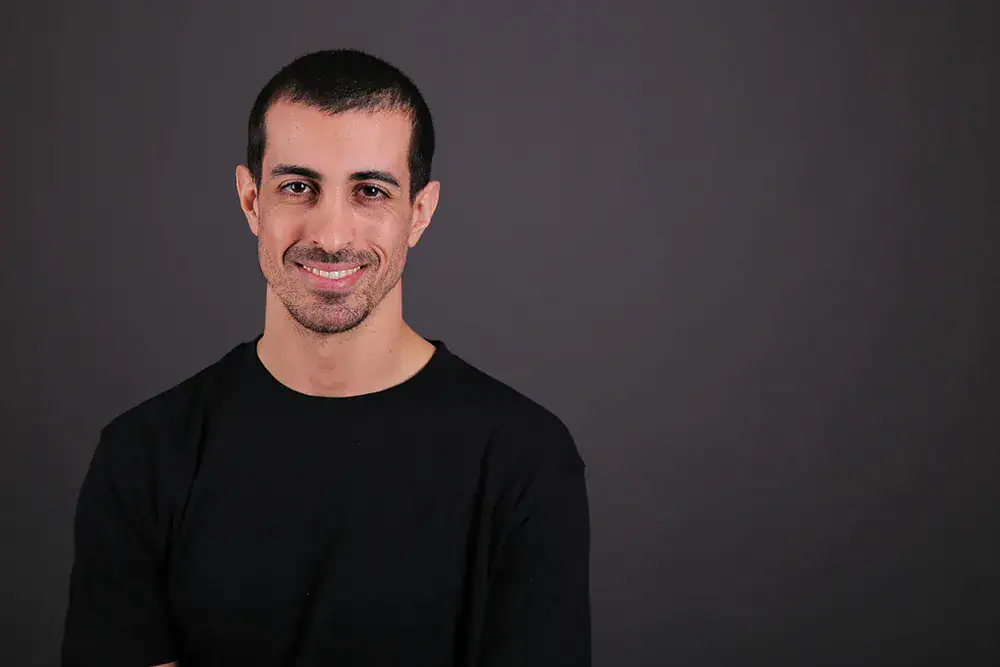 Headshot example featuring a smiling person in a plain black shirt, positioned off-center against a dark gray studio backdrop. The minimalist styling and relaxed posture create a warm, approachable, modern-professional impression.
