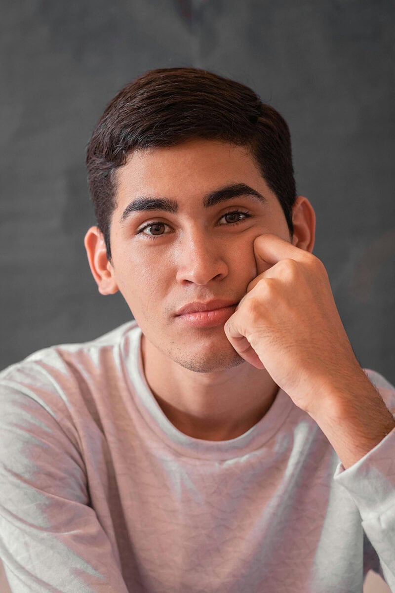 Headshot example featuring a young man in a relaxed, hand-to-face pose with an introspective expression, set against a dark, textured gray background with intimate tight framing.