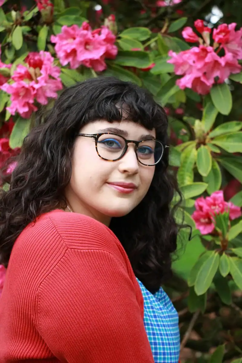 Headshot example featuring a creative professional with tortoiseshell glasses and blue eyeliner, posed against a lush backdrop of vibrant pink floral blossoms and green foliage.