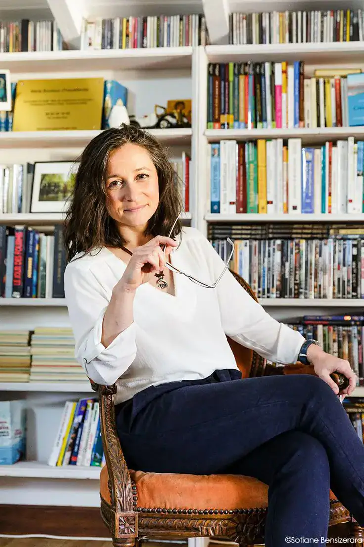 Headshot example featuring a person seated in front of tall bookshelves filled with books, holding a pair of glasses near their face. Natural light, relaxed posture, and the library setting create an intellectual, thoughtful, welcoming mood.