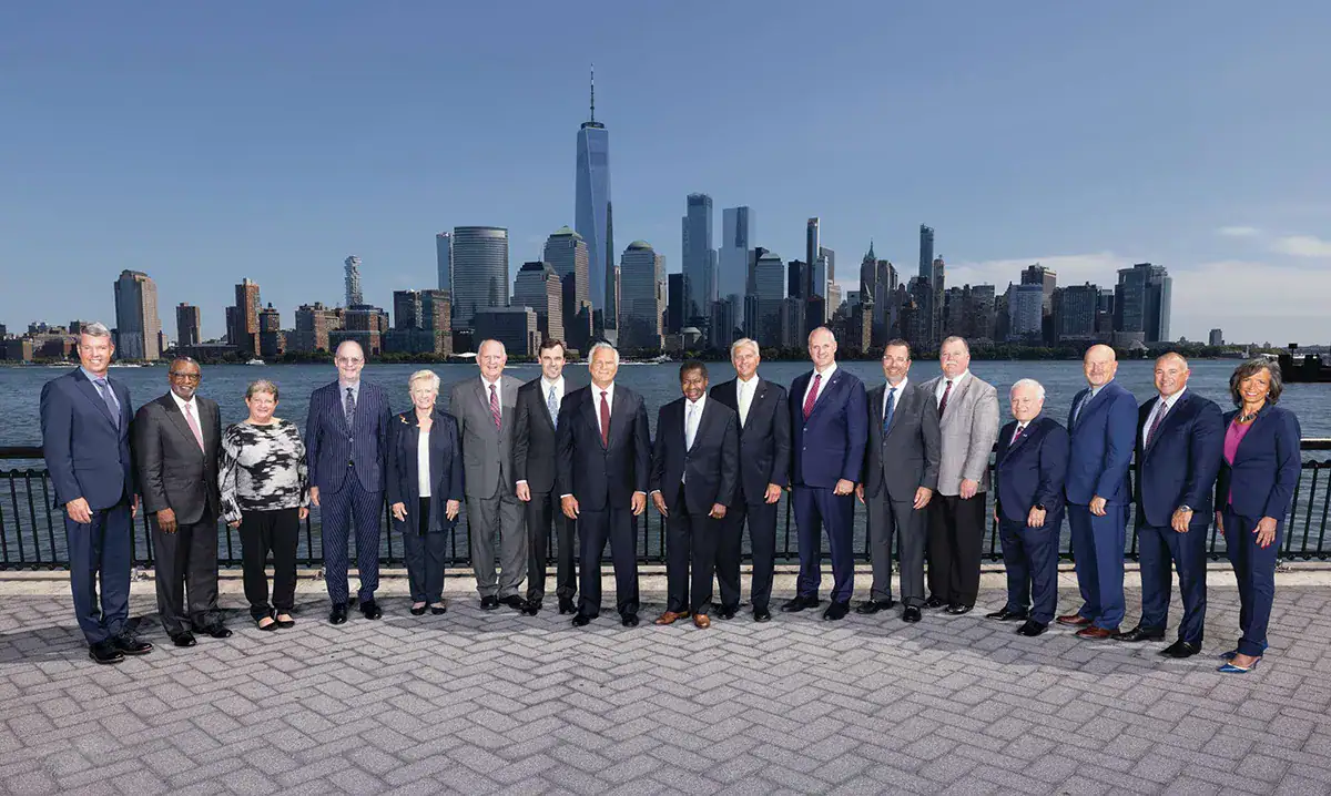 A group of 17 professionally dressed men and women stand in a long line on a paved waterfront promenade for an environmental portrait in NYC. In the background, the bright blue sky meets the extensive Lower Manhattan skyline, featuring One World Trade Center prominently in the center above the Hudson River.