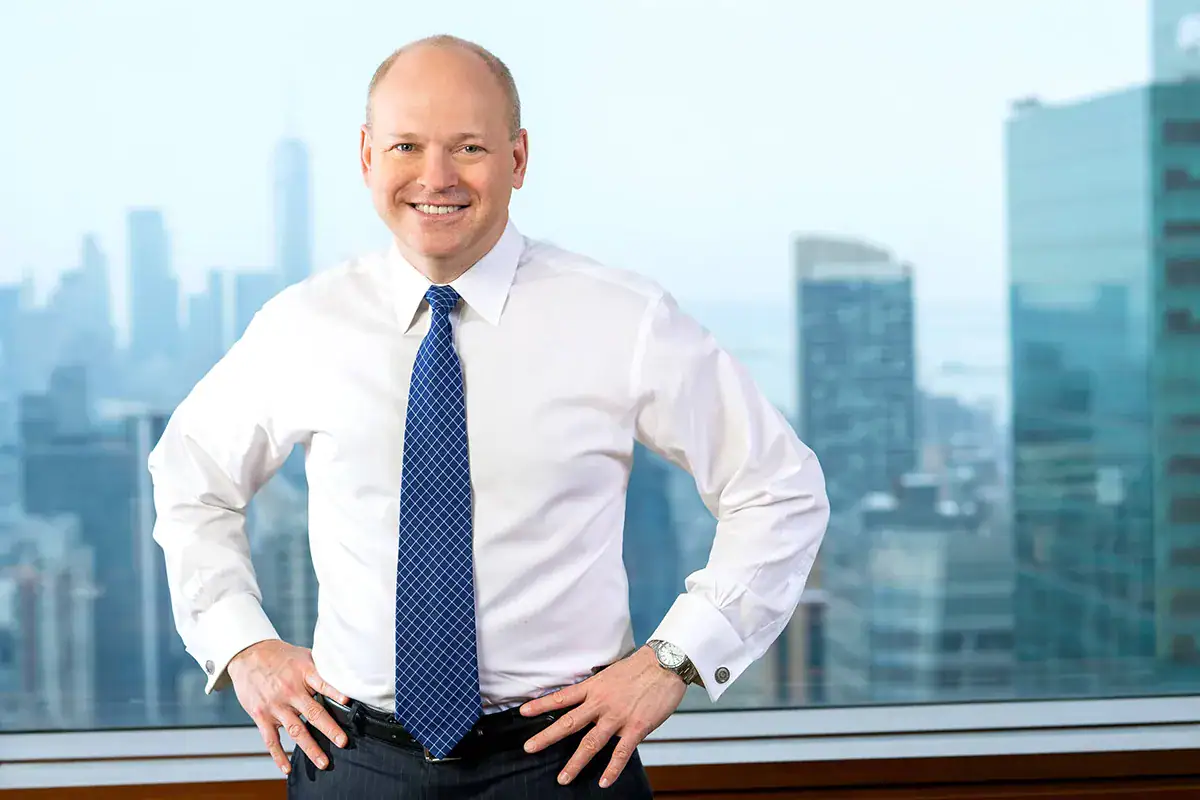 A professional man in a white dress shirt and blue tie smiles with his hands on his hips during an environmental headshot portrait in NYC. He stands indoors in front of a high-rise office window overlooking a blurred Manhattan skyline with skyscrapers visible in the distance.