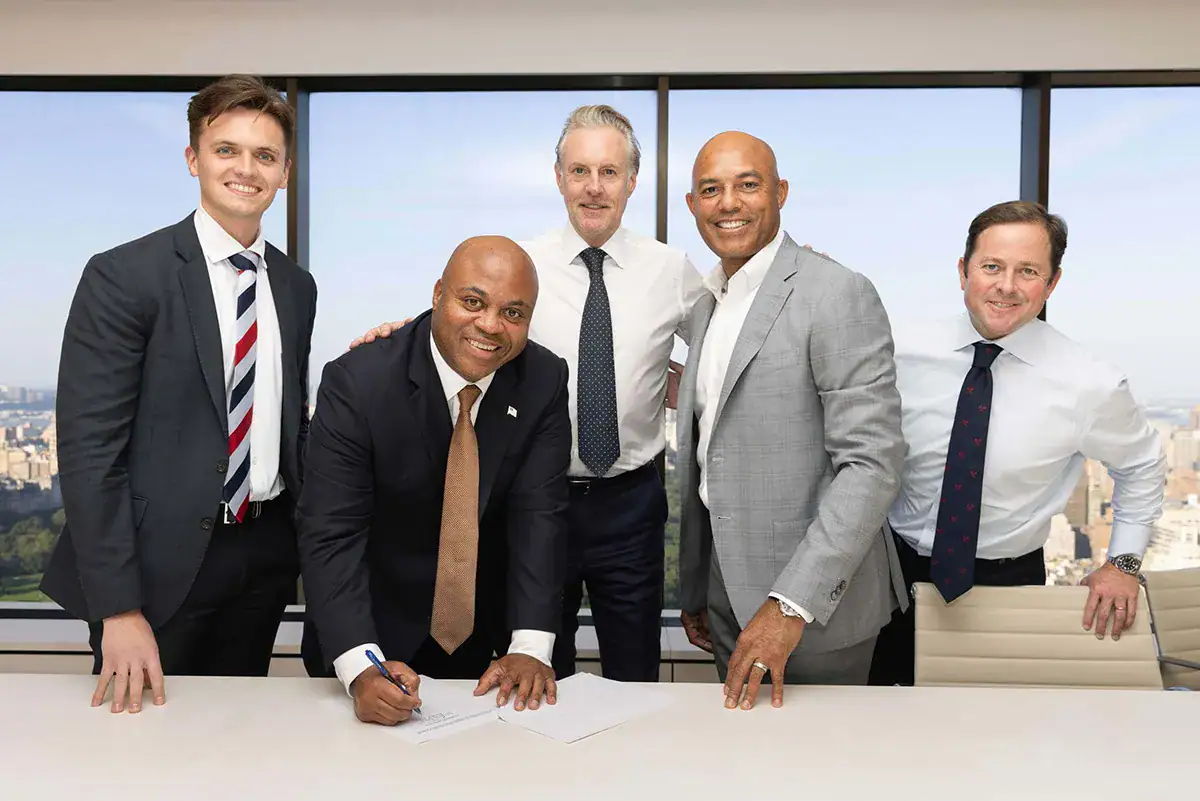 Five professionally dressed men gather around a white conference table during a document signing inside a high-rise NYC office. One man leans forward to sign a paper, while the others stand alongside him smiling. A large window in the background overlooks a high-angle view of the Manhattan skyline and Central Park.