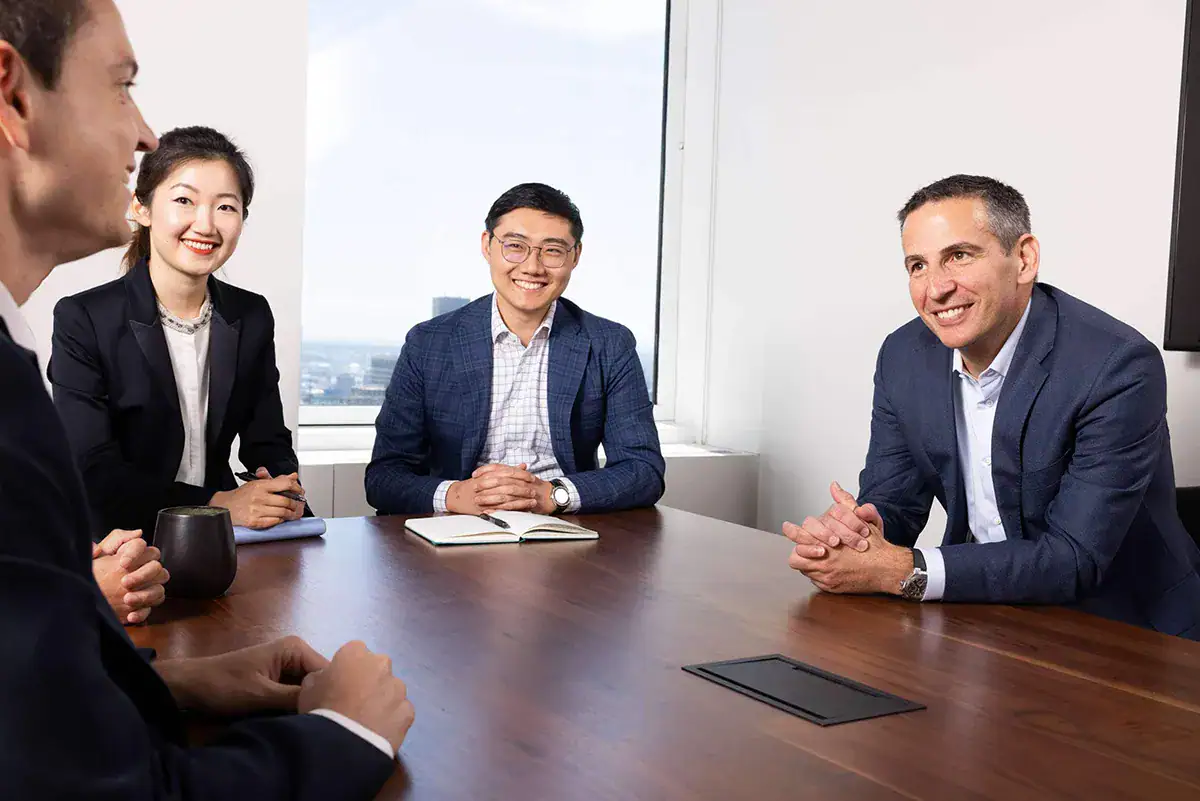 Four corporate professionals sit around a dark wood conference table during a candid meeting inside an NYC high-rise office. Three individuals smile while interacting with a colleague whose profile is visible in the foreground. A large window in the background shows a bright view of a distant city skyline under a clear sky.
