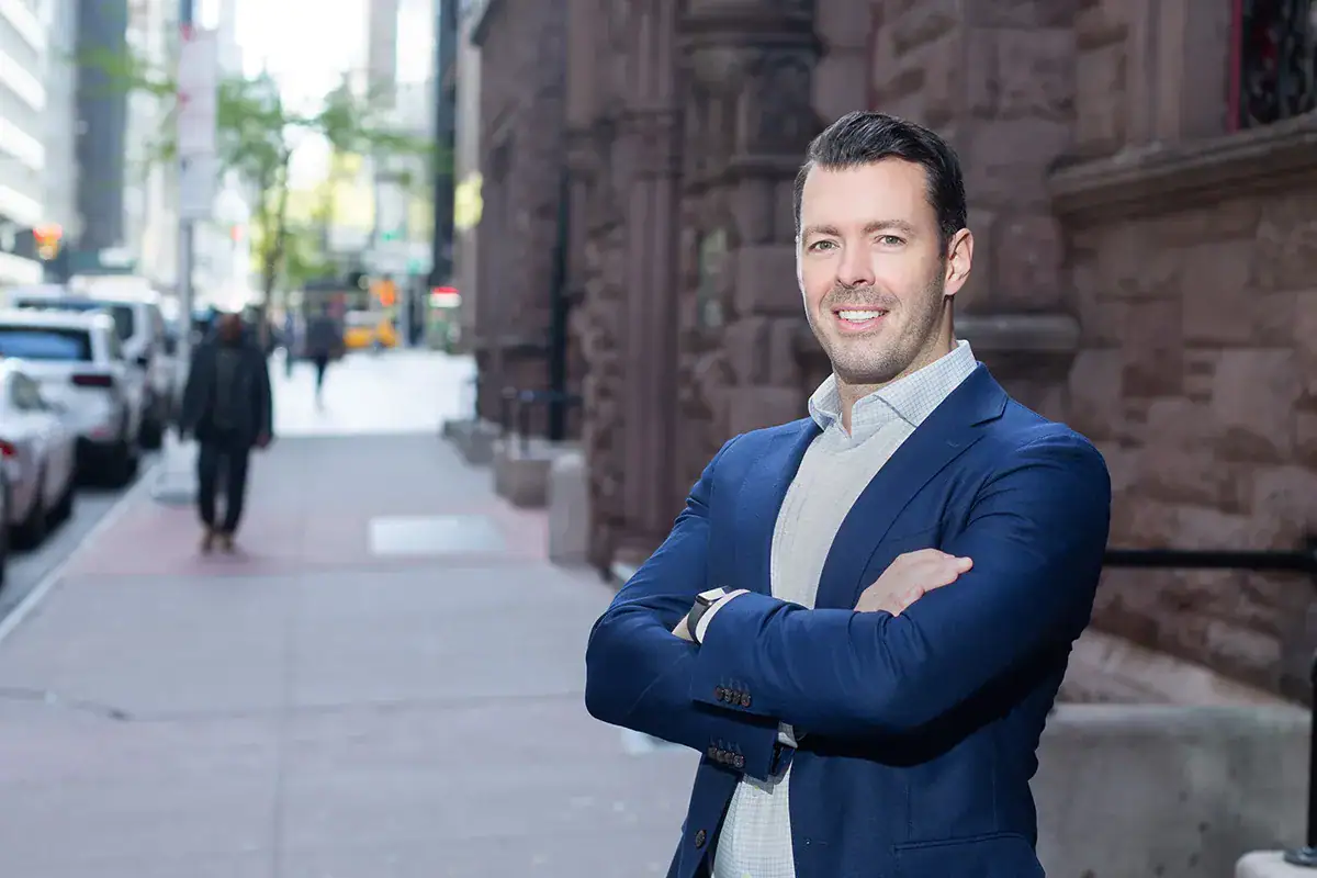 A professional man in a blue blazer and light patterned dress shirt stands with his arms crossed and smiles during an outdoor portrait on a Manhattan sidewalk. He stands next to a classic stone building facade, with a blurred street background showing parked cars, a pedestrian, and city elements extending down the block.