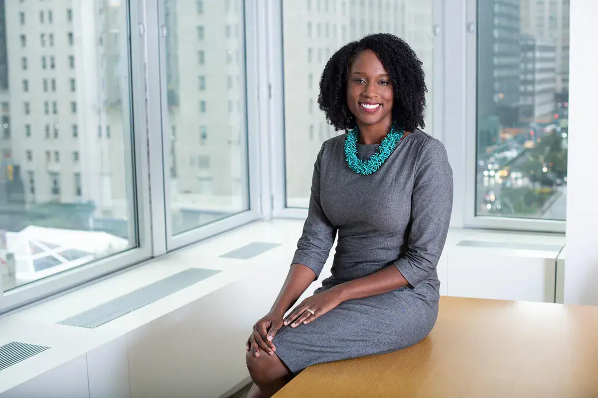 A professional woman with dark curly hair, wearing a grey dress and a turquoise necklace, sits on the edge of a light wood desk and smiles during an indoor portrait. Large windows wrap around the corner office, showcasing a blurred background of NYC skyscrapers and a street below.