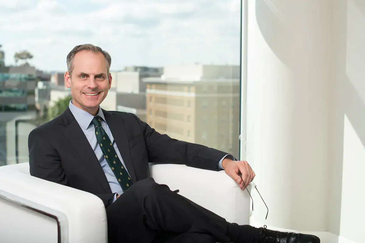 A professional man in a dark suit, light blue dress shirt, and green patterned tie sits with his legs crossed in a white armchair while holding eyeglasses and smiling. A large window behind him showcases a blurred view of NYC buildings under a cloudy sky.