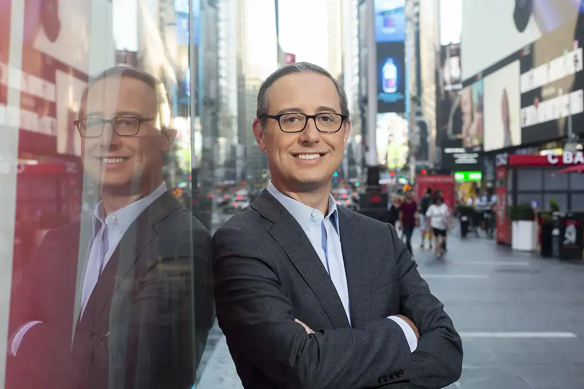 A professional man wearing glasses, a dark grey suit jacket, and a light blue dress shirt stands with arms crossed and smiles next to a reflective glass wall that captures his reflection. The blurred background features the large digital billboards, towering buildings, and pedestrians of Times Square in NYC.