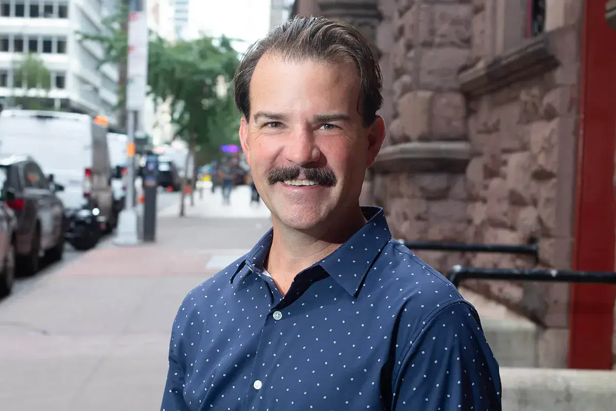 A man with a mustache wearing a blue polka-dot button-down shirt smiles during an outdoor headshot on an NYC sidewalk. He stands next to a textured stone building facade on 46th Street, with a blurred background showing parked vehicles, a tree, and street elements extending down the block.