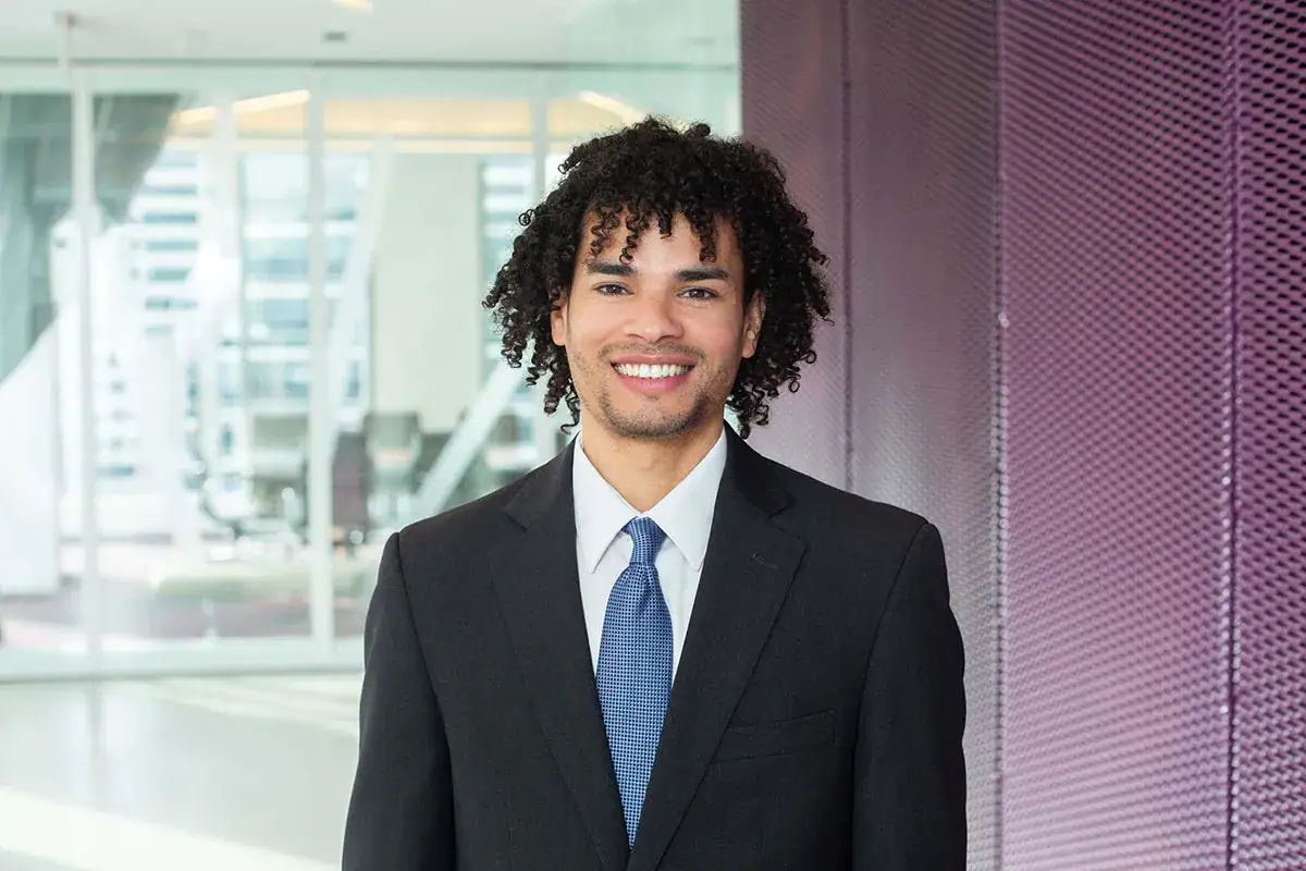 A professional young man with curly dark hair and a smile stands during a corporate headshot in an NYC office. He wears a dark suit jacket, white dress shirt, and a blue textured tie, positioned next to a purple mesh accent wall with a blurred modern office interior in the background.