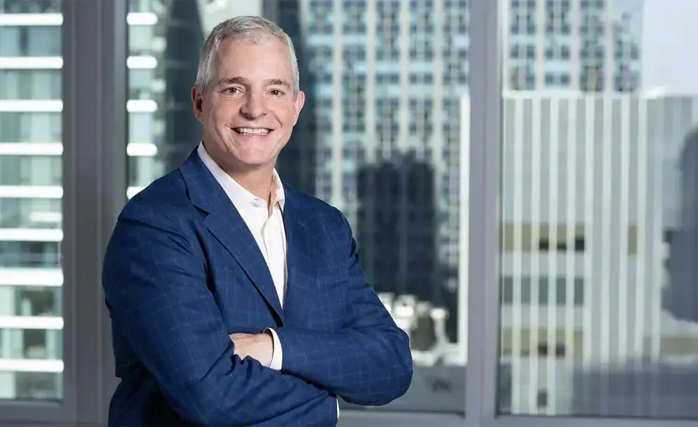 A professional man with short gray hair and a warm smile stands with his arms crossed during a corporate headshot. He wears a blue checked blazer and a white dress shirt, positioned tightly against large office windows that completely frame him with a blurred backdrop of towering NYC skyscrapers.