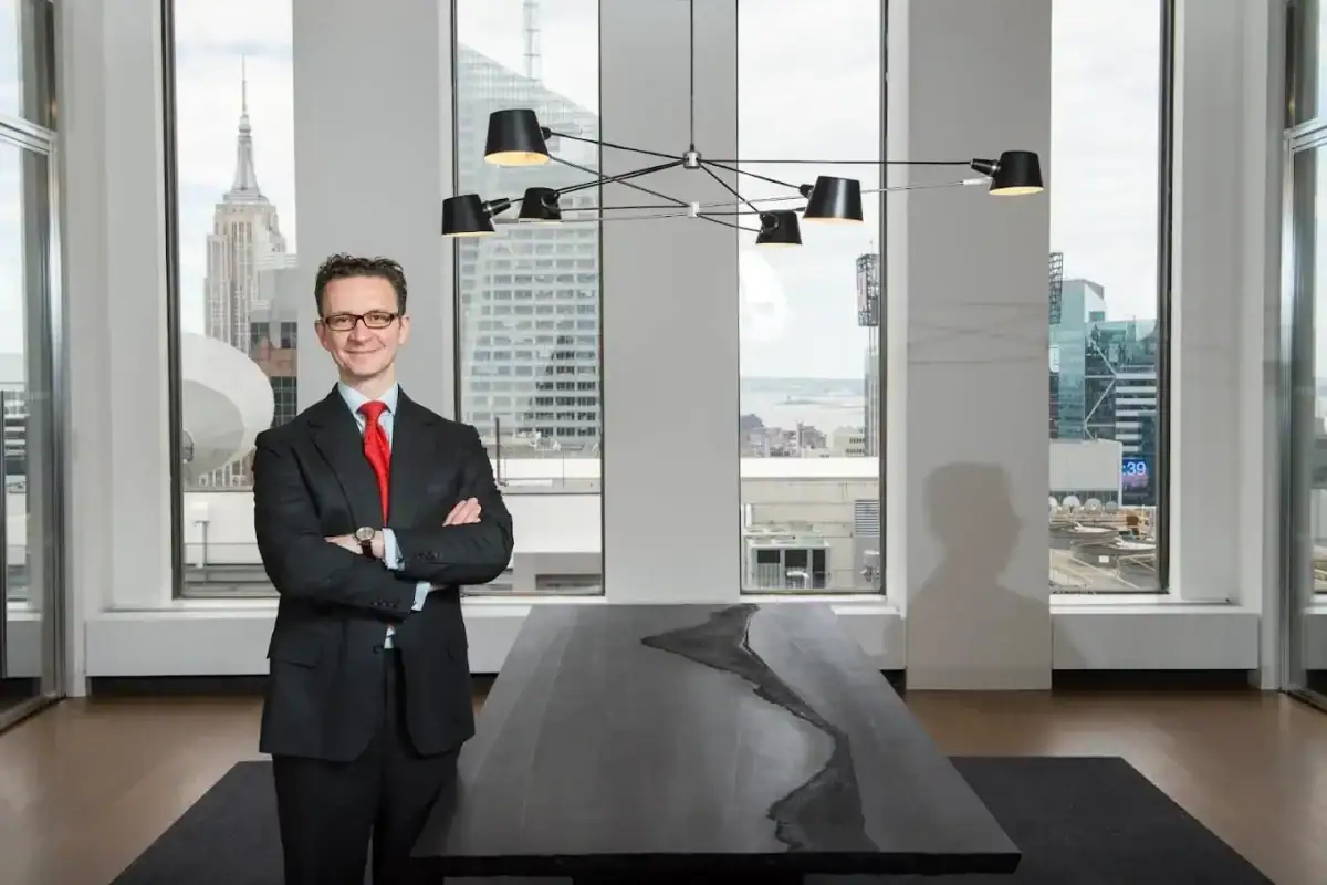 A professional man with glasses and a smile stands with his arms crossed in a modern NYC boardroom. He is wearing a dark suit and a vibrant red tie, positioned to the left of a large, dark conference table. Behind him, tall vertical windows separated by white columns showcase a panoramic high-rise city view featuring the Empire State Building.