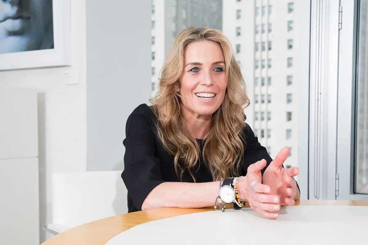 A professional woman with wavy blonde hair smiles and gestures with her hands during a candid, high-key portrait in a modern office. She wears a black mid-sleeve top and a white-faced watch, seated at a light-colored round table. The bright background showcases large windows with a softly blurred metropolitan view and a framed black-and-white artwork on the wall.