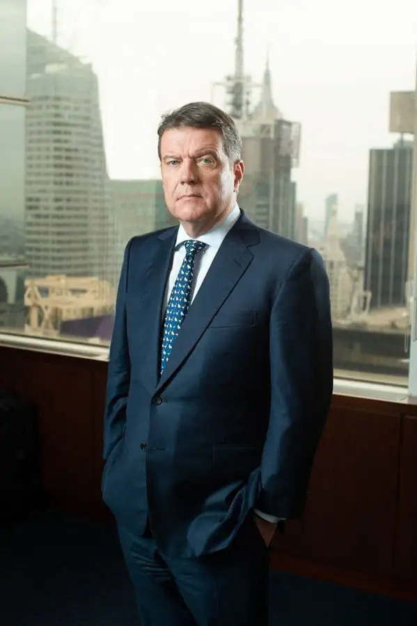 A professional man with a serious expression stands with his hands in his pockets during a corporate portrait. He wears a dark blue suit and a patterned tie, positioned before large office windows that showcase a blurred Midtown Manhattan skyline with prominent skyscrapers.