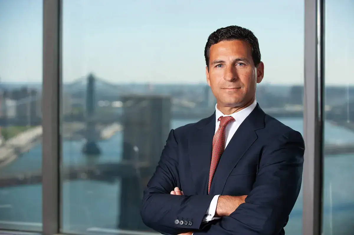 A professional man with short dark hair stands with crossed arms in a corporate headshot. He wears a dark blue suit, a white dress shirt, and a red patterned tie. Behind him, large windows provide a bright, high-rise view of the East River, showcasing the Brooklyn Bridge and the Manhattan Bridge in a soft-focus background.