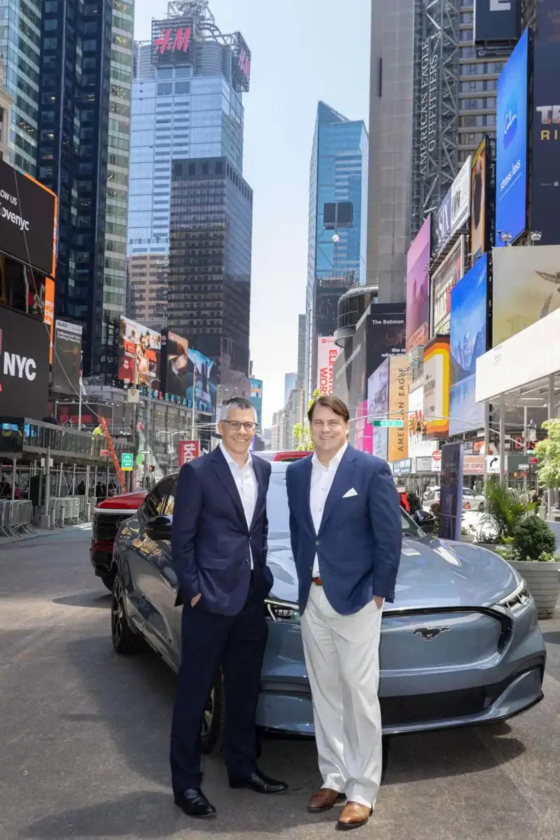 Two men in business attire stand in front of a grey Ford Mustang Mach-E parked in the middle of Times Square, New York City. The man on the left wears a dark suit with glasses, while the man on the right wears a blue blazer and light trousers. They are surrounded by towering skyscrapers and vibrant digital billboards under a bright sky.