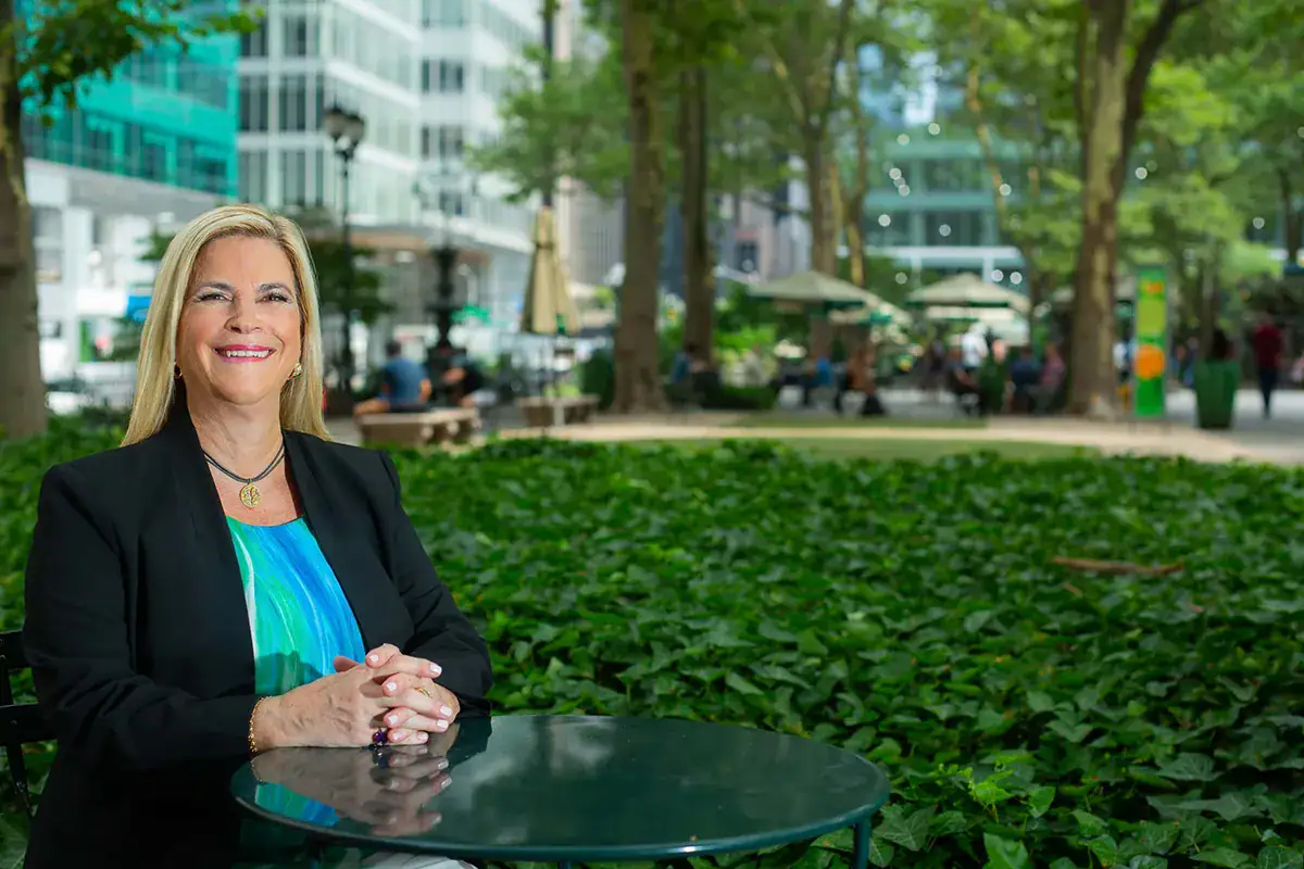 A professional woman with blonde hair sits smiling at a green round table in an outdoor park. She wears a black blazer over a blue and green patterned top, with her hands clasped on the table. The background features a thick bed of green ivy, mature trees, park seating with umbrellas, and blurred urban buildings.