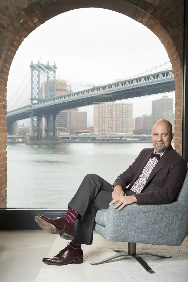 A professional man with a beard sits smiling in a grey armchair, framed by a large, distinctive arched brick window. He wears a dark blazer, checkered shirt, bow tie, grey trousers, and burgundy dress shoes with matching striped socks. Through the glass, the Brooklyn Bridge spans across the water against a city backdrop under an overcast sky.