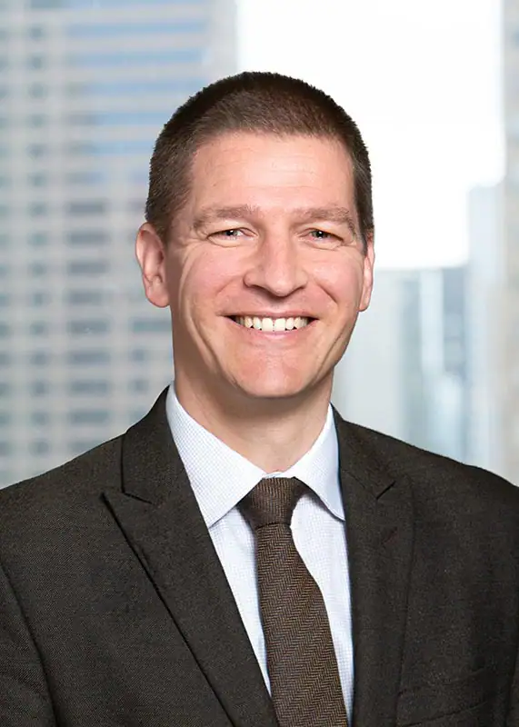 A smiling man with short brown hair in a close-up corporate headshot. He wears a dark, textured suit jacket, a light checkered dress shirt, and a brown patterned tie. The background is a blurred NYC high-rise view of city buildings through an office window.
