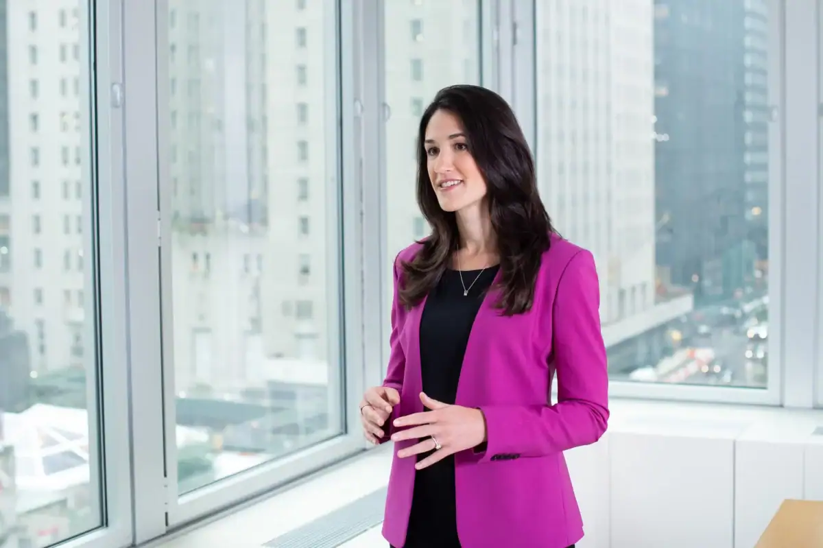 A professional woman with long dark hair stands smiling and gesturing next to large office windows. She wears a vibrant fuchsia blazer over a black dress. The background features a soft-focus, high-rise view of city skyscrapers through the glass.